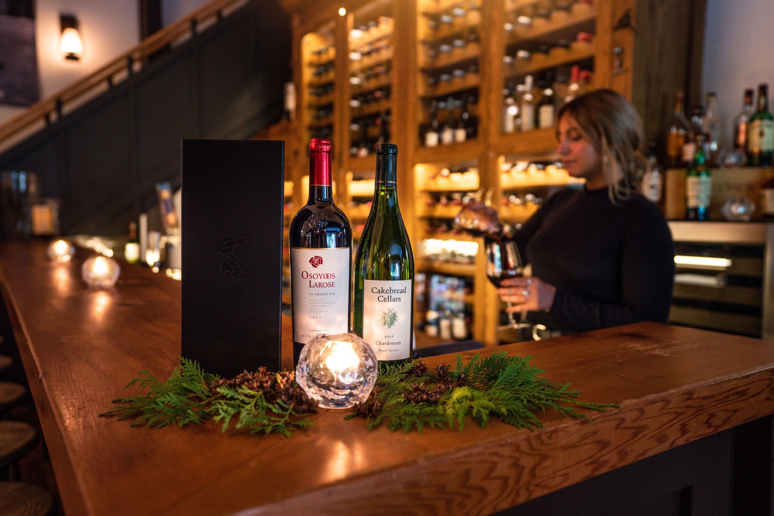 Behind the bar at Tweedsmuir Split Rock Restaurant, a woman poses with several wine bottles displayed on the counter.